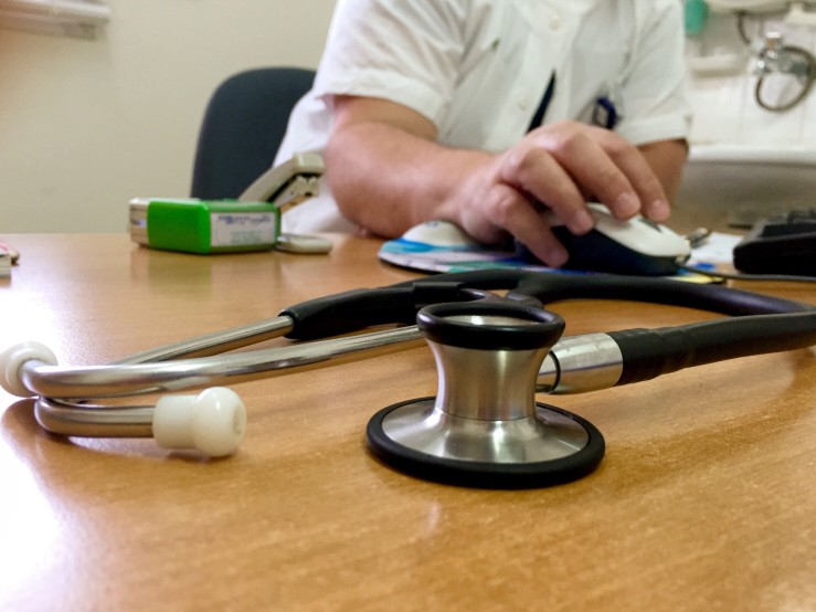 A doctor's desk with a stethoscope in the foreground and a doctor writing in on a pad in the background