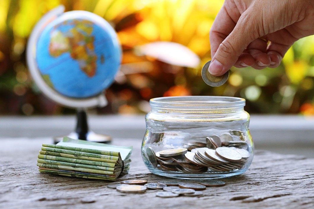 People putting coins in glass jar and money bank with a blurry world globe map closeup in the background